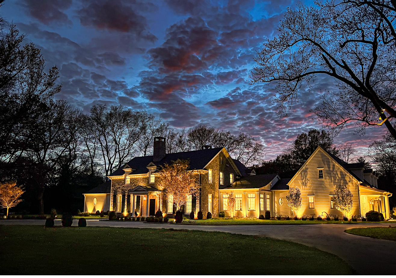 Grand stone estate with full architectural lighting package under dramatic sunset sky